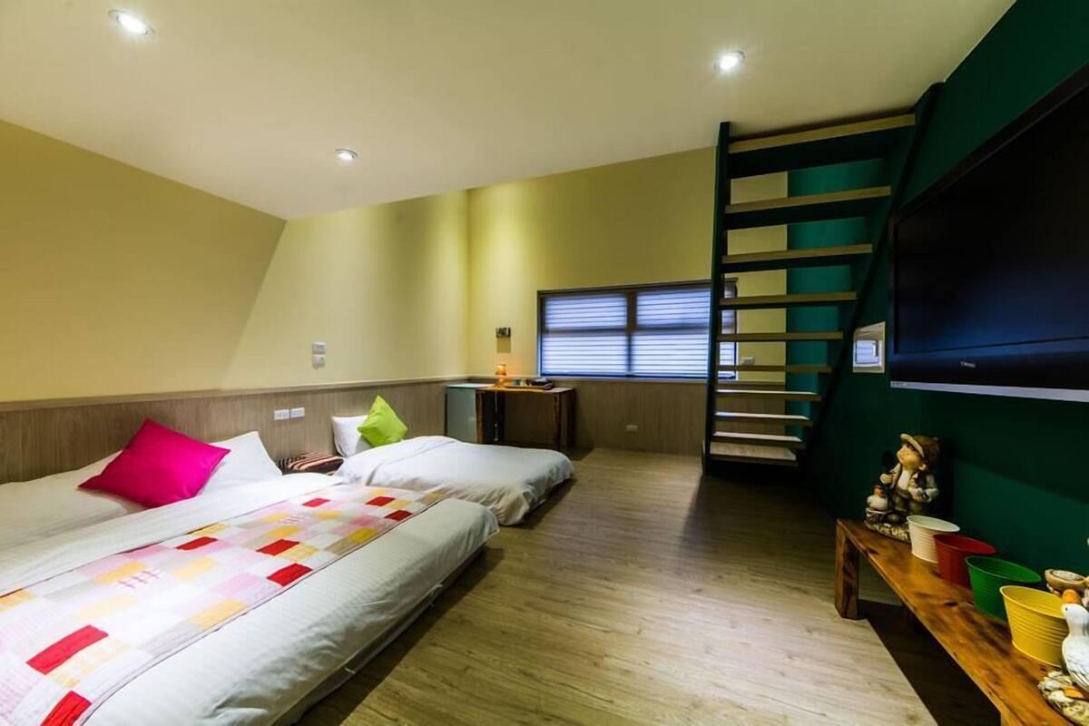 A bedroom with two beds, a wooden floor, a window with blinds, a small table with a lamp, and a green wall.