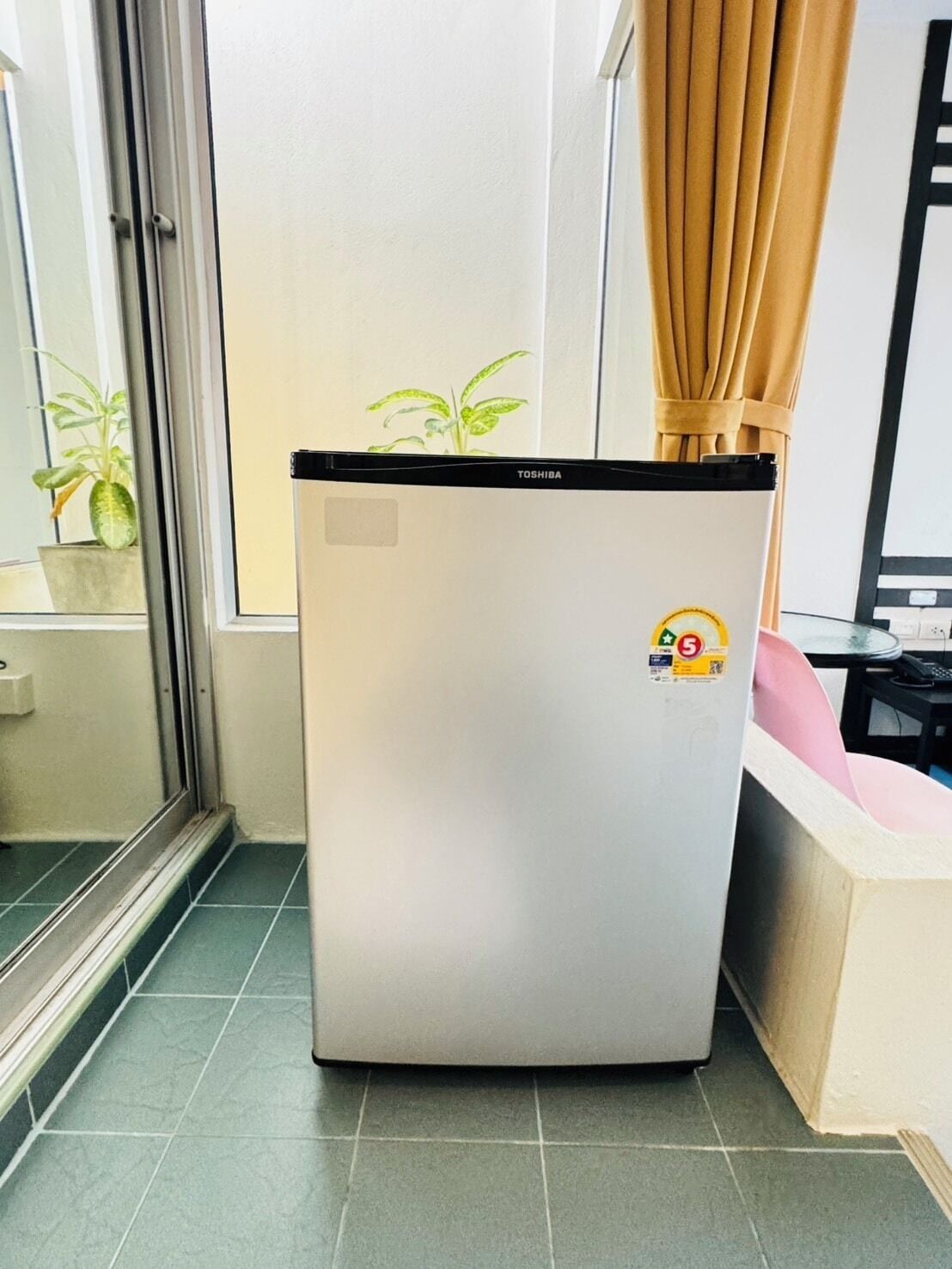 A compact refrigerator in a room with a sliding glass door, a potted plant, and a pink chair.