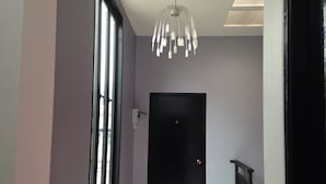 Um corredor com uma porta preta, uma janela com persianas e um lustre moderno.