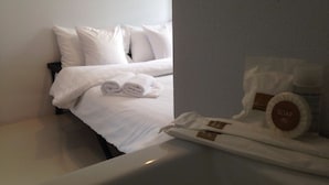 Cama bem arrumada com lençóis brancos e toalhas dobradas, uma mesinha com artigos de higiene pessoal, incluindo sabonete e loção, e uma parede lisa ao fundo.