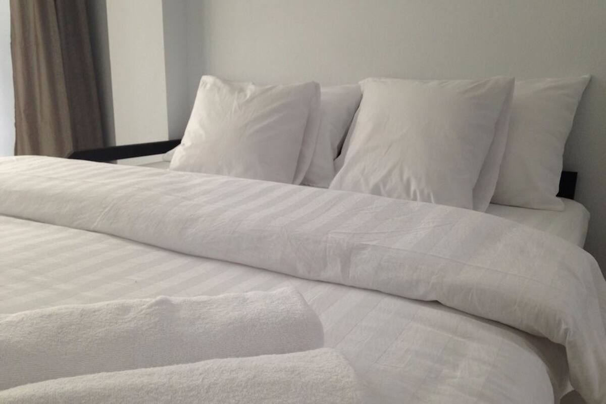 A neatly made bed with white bedding and pillows, a window with curtains, and a plain wall.