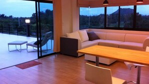 A modern living room with a sofa, a dining table, and a chair. There is a large window with a view of a mountain and trees.