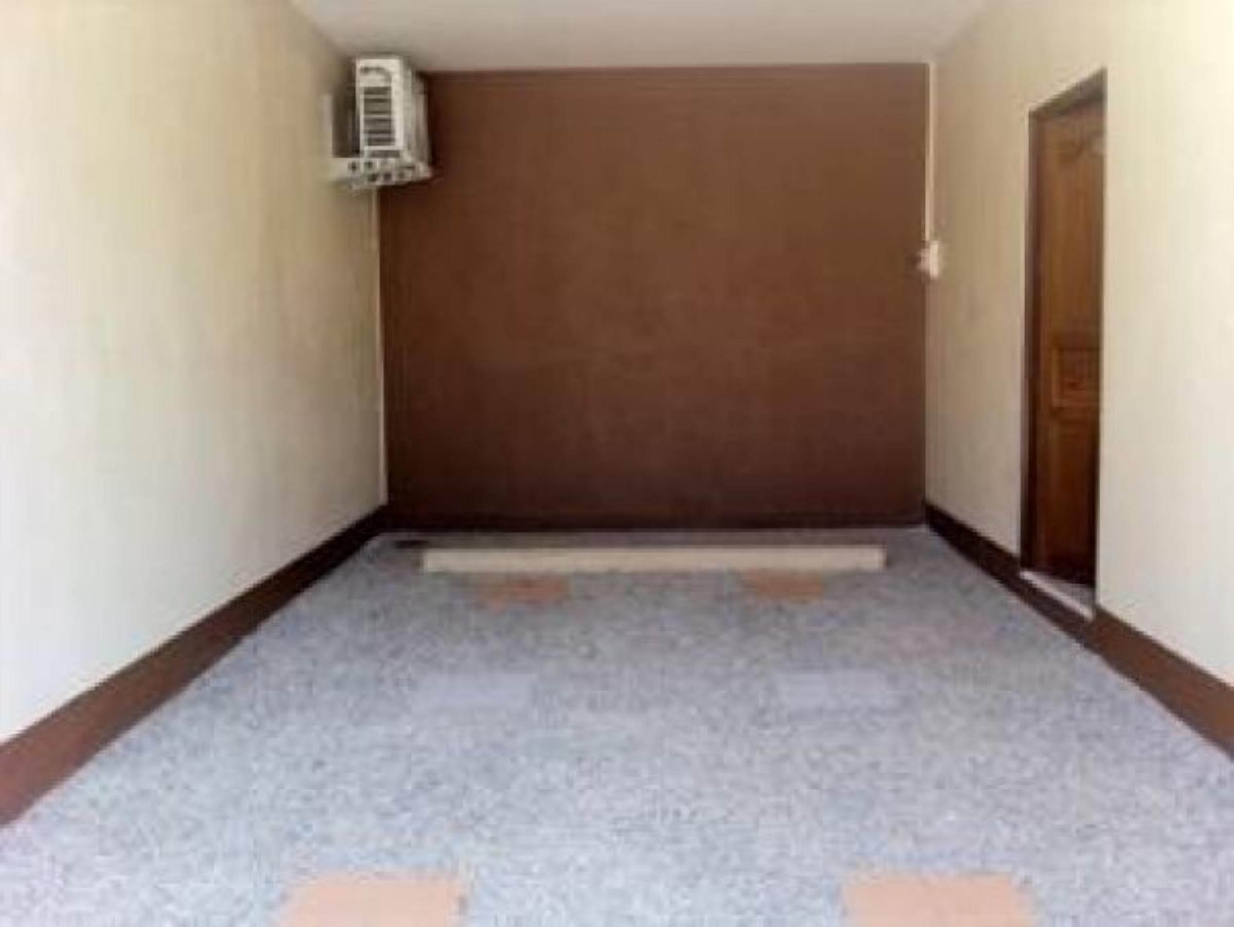 Un pasillo estrecho con una puerta marrón al final, un equipo de aire acondicionado blanco en la pared izquierda y una alfombra gris.