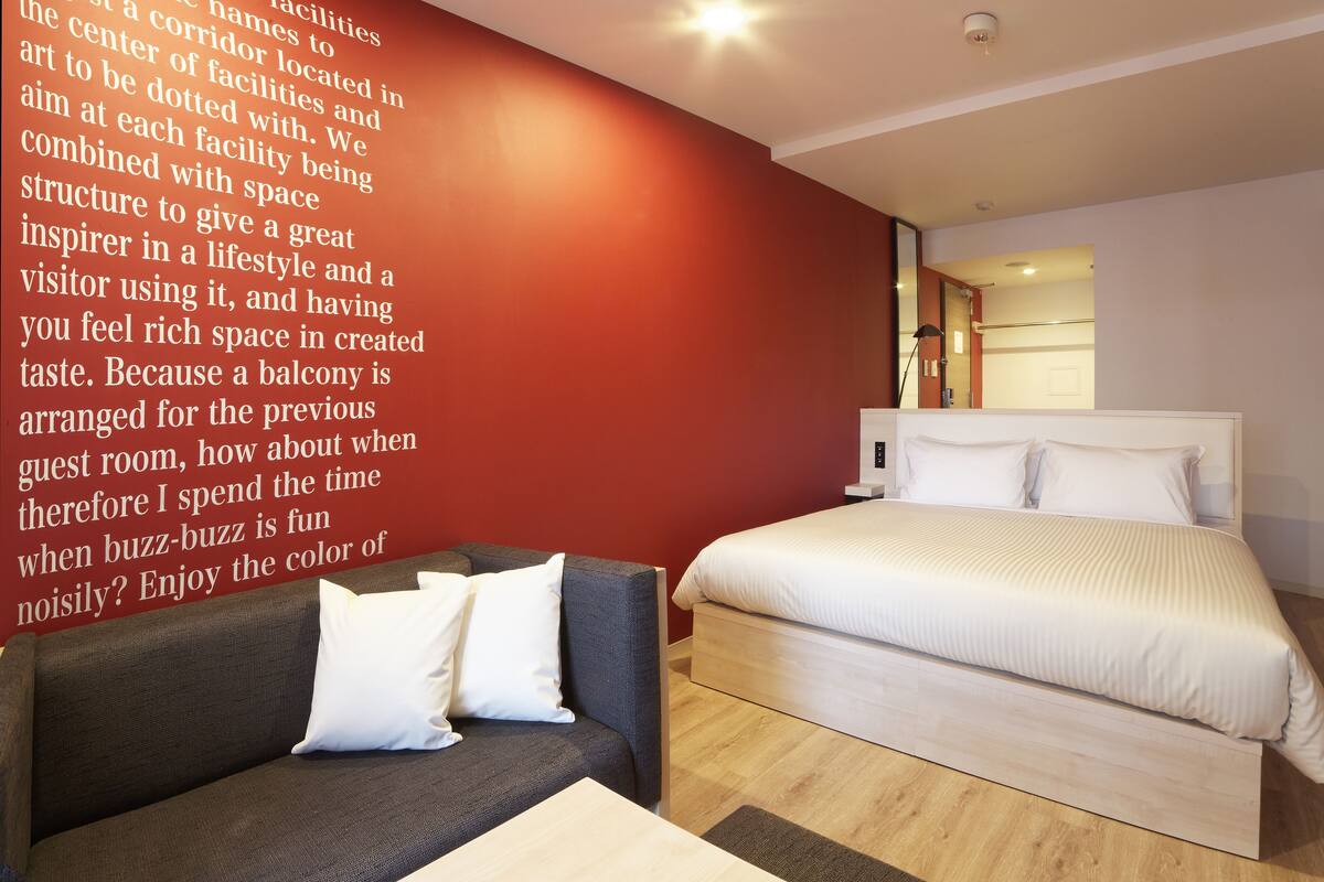 A modern hotel room with a large bed, a sofa, and a red accent wall.