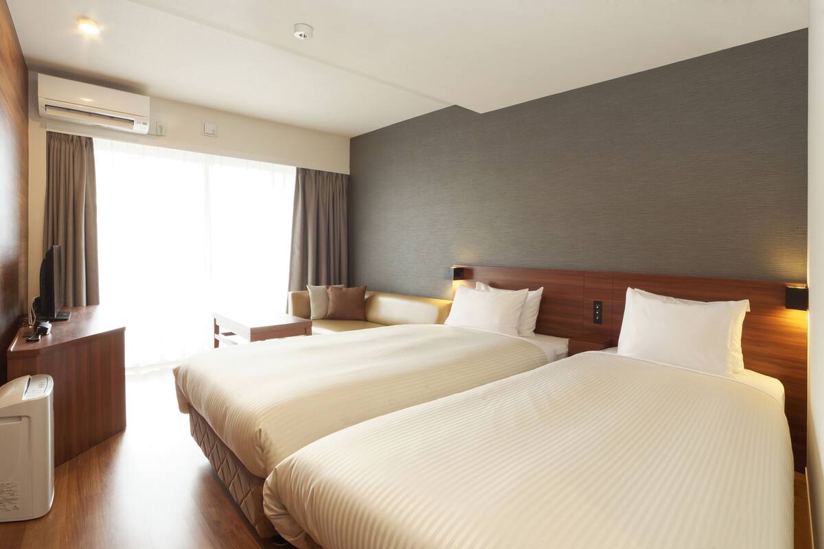 A hotel room with two beds, a desk, an air conditioning unit, and a view of the outdoors.