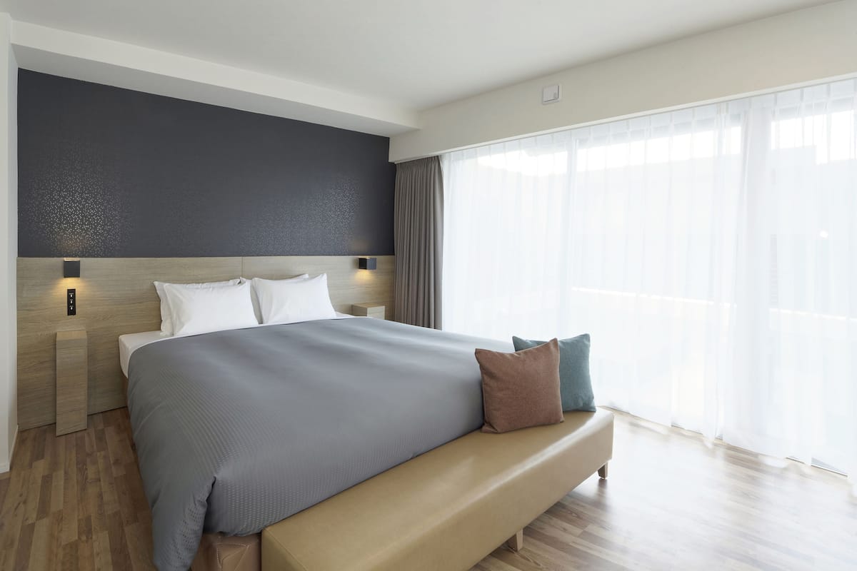 A modern hotel room with a large bed, a bench, and a view of the city through sheer curtains.