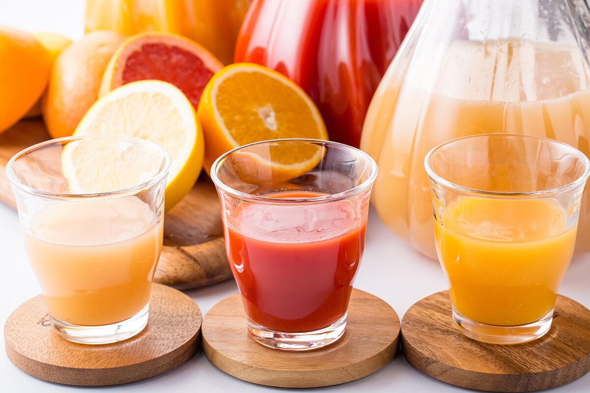 Three glasses of juice on wooden coasters, with a pitcher and citrus fruits in the background.