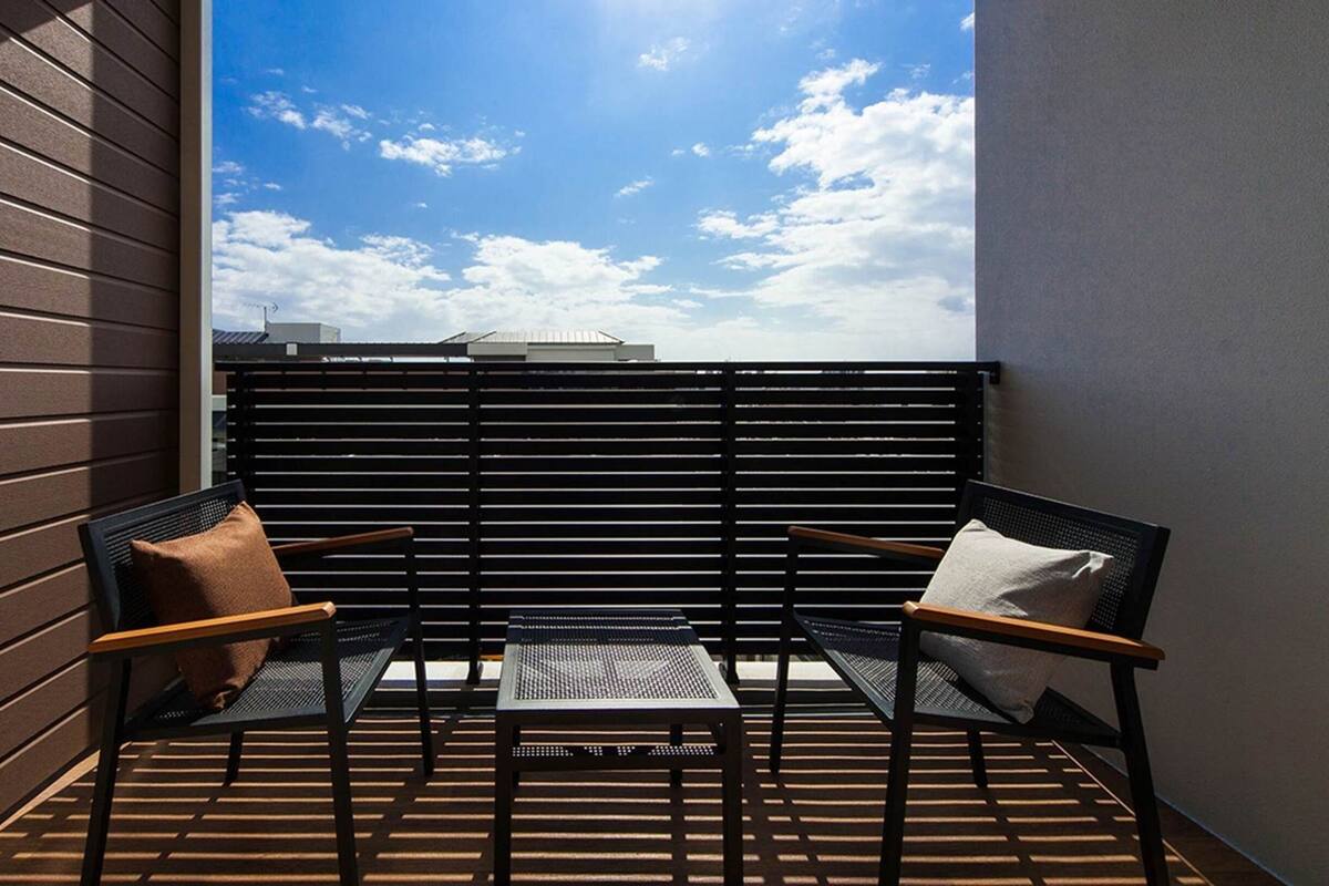 A small balcony with two chairs and a table, a slatted black fence, and a view of the sky and buildings.