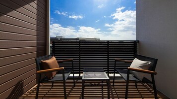 A small balcony with two chairs and a table, a slatted black fence, and a view of the sky and buildings.