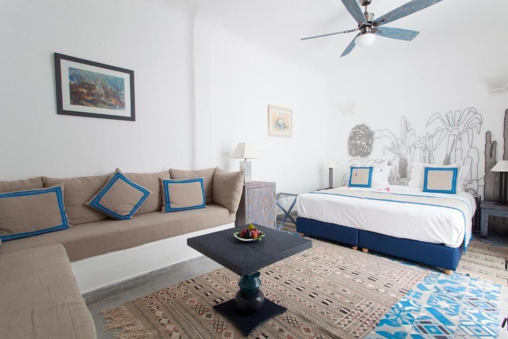 Un dormitorio con una cama, un sofá, una mesita con fruta, un ventilador de techo y una pared con una ilustración de una planta y un cactus.