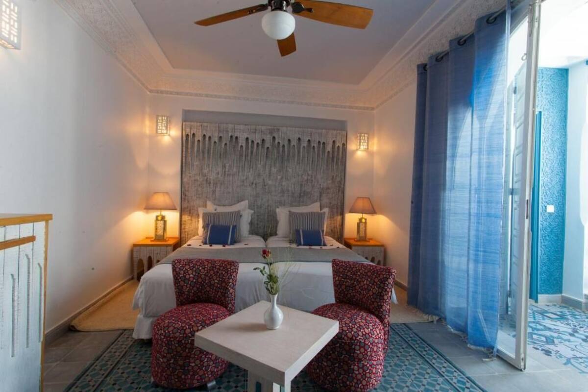 A hotel room with a bed, two bedside tables, a ceiling fan, a table with a vase, and patterned chairs.