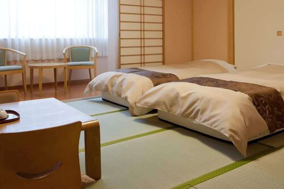 A hotel room with a bed, a chair, a table, and a window with blinds.