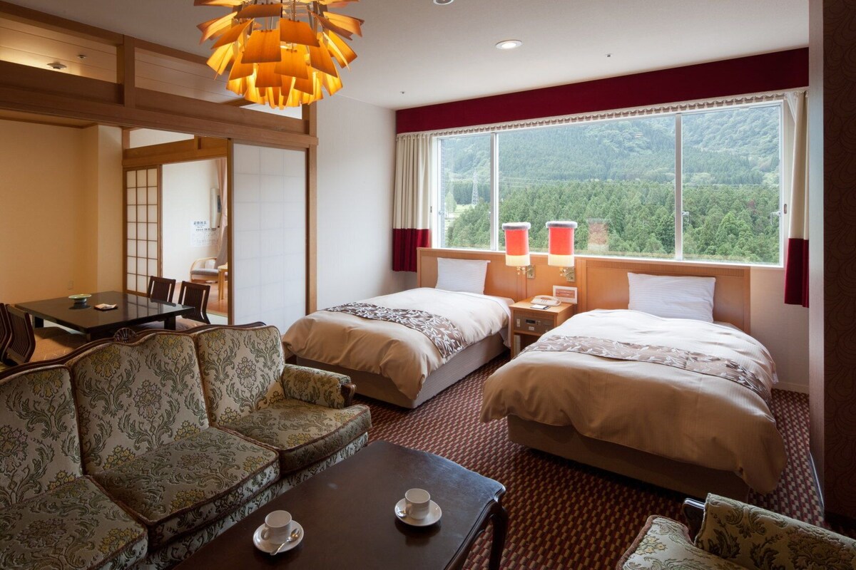 A hotel room with a sofa, two beds, a dining table, and a view of the forest.