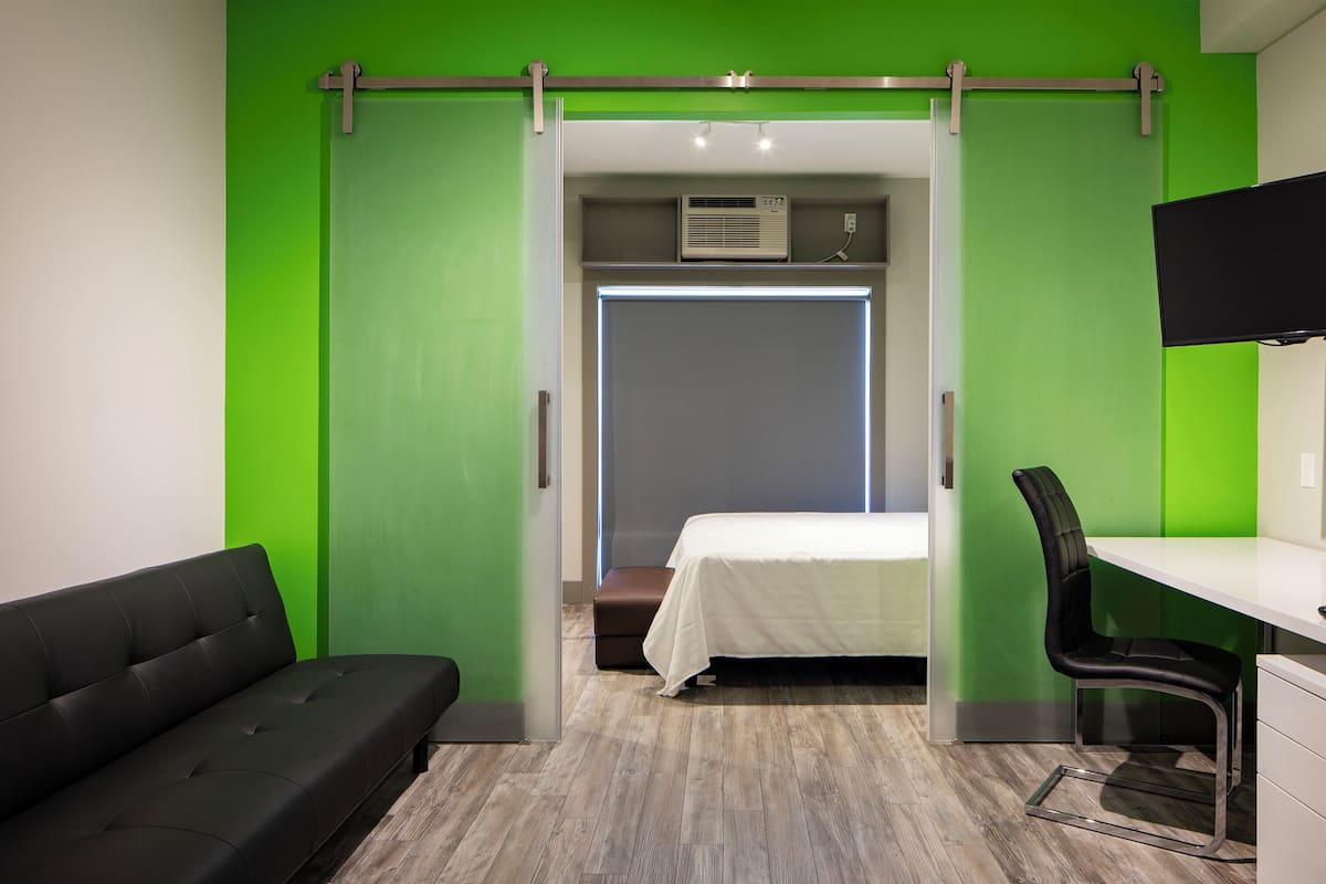 A modern hotel room with a green accent wall, a bed, a desk with a chair, and a sofa.