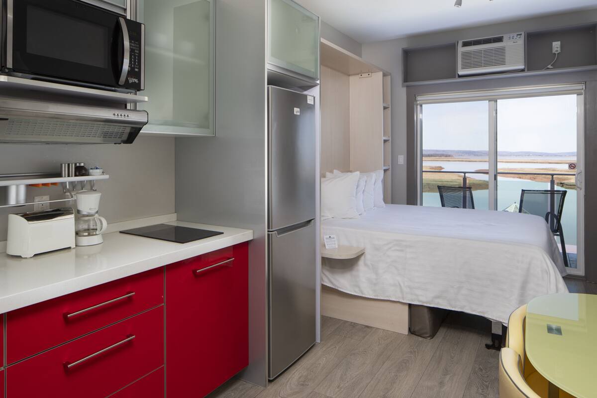 A modern hotel room with a kitchenette, a bed, a balcony with a view, and a glass table.