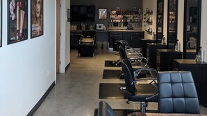 Un salón de belleza con varias sillas de peluquería, un mostrador de recepción y estanterías llenas de productos capilares.