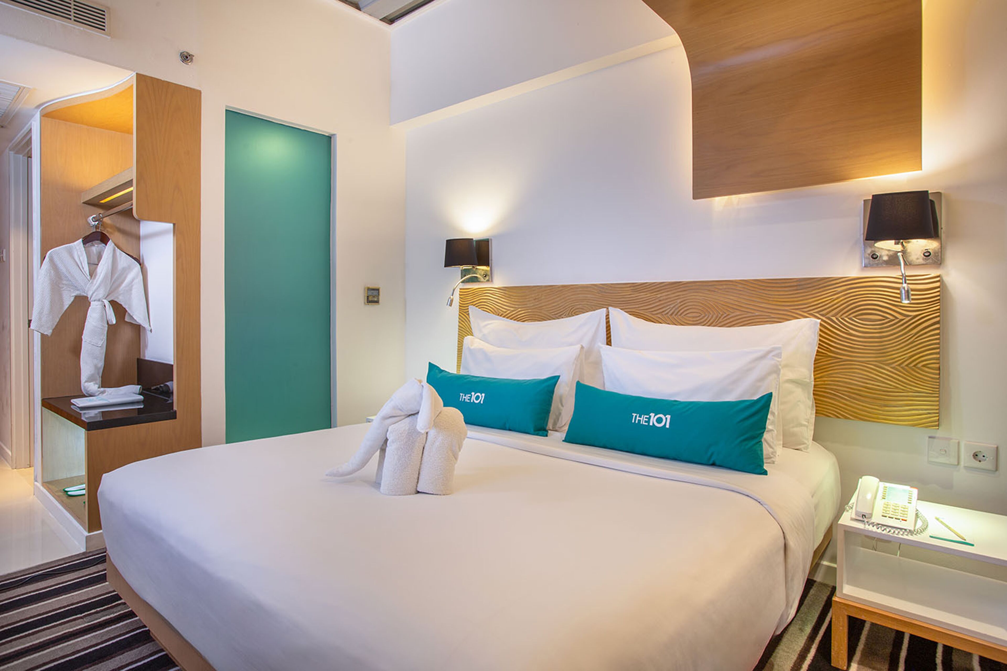 Een hotelkamer met een opgemaakt bed, een nachtkastje met een telefoon en notitieboekje, en een wandlamp.