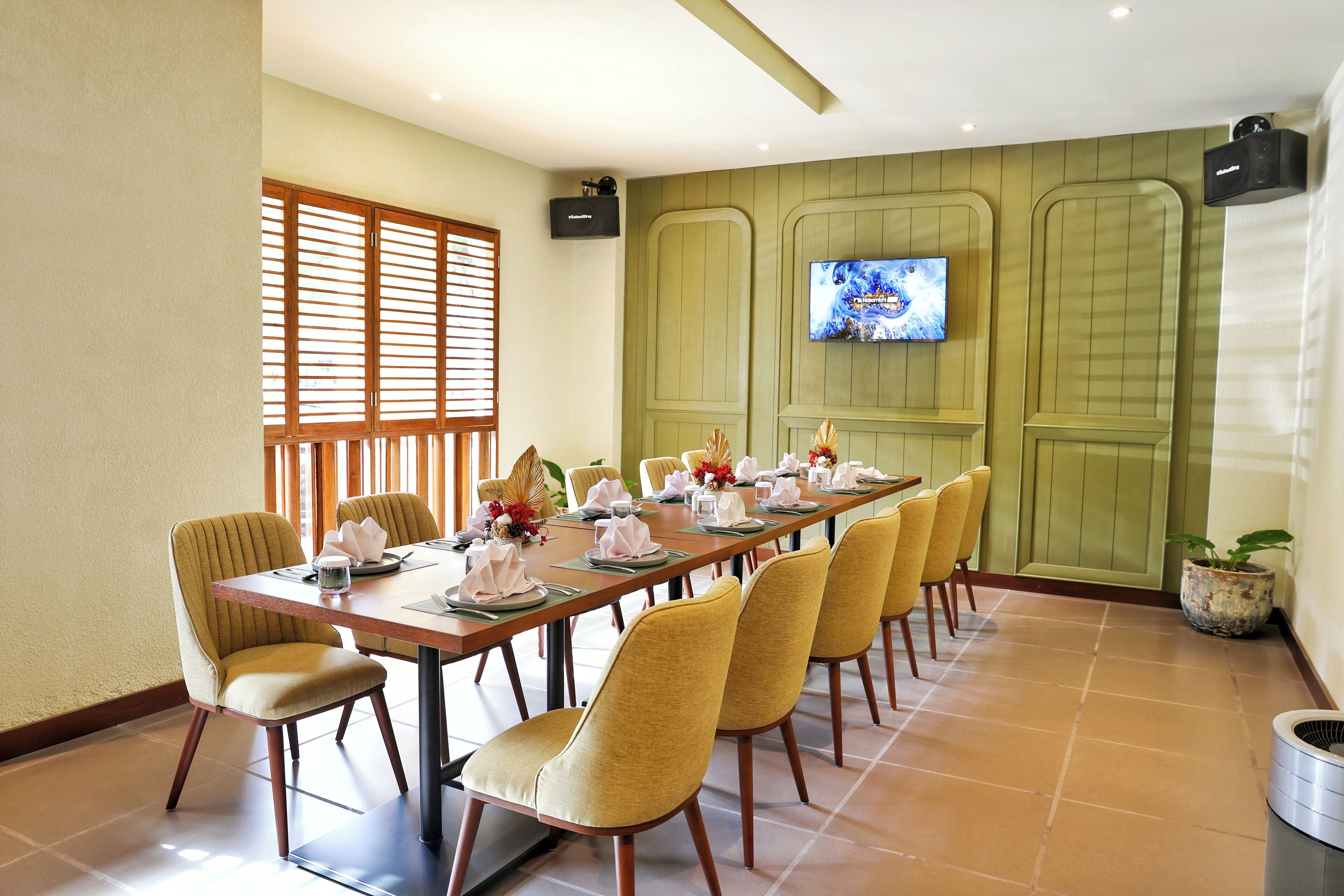 A dining area with a long table set for a meal, surrounded by yellow chairs, with a television mounted on the wall and a window with blinds.