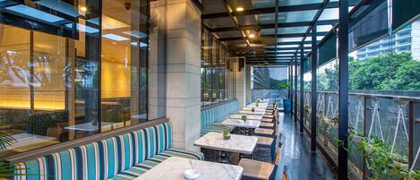 A covered outdoor seating area with striped benches and tables, large windows, and greenery visible outside.