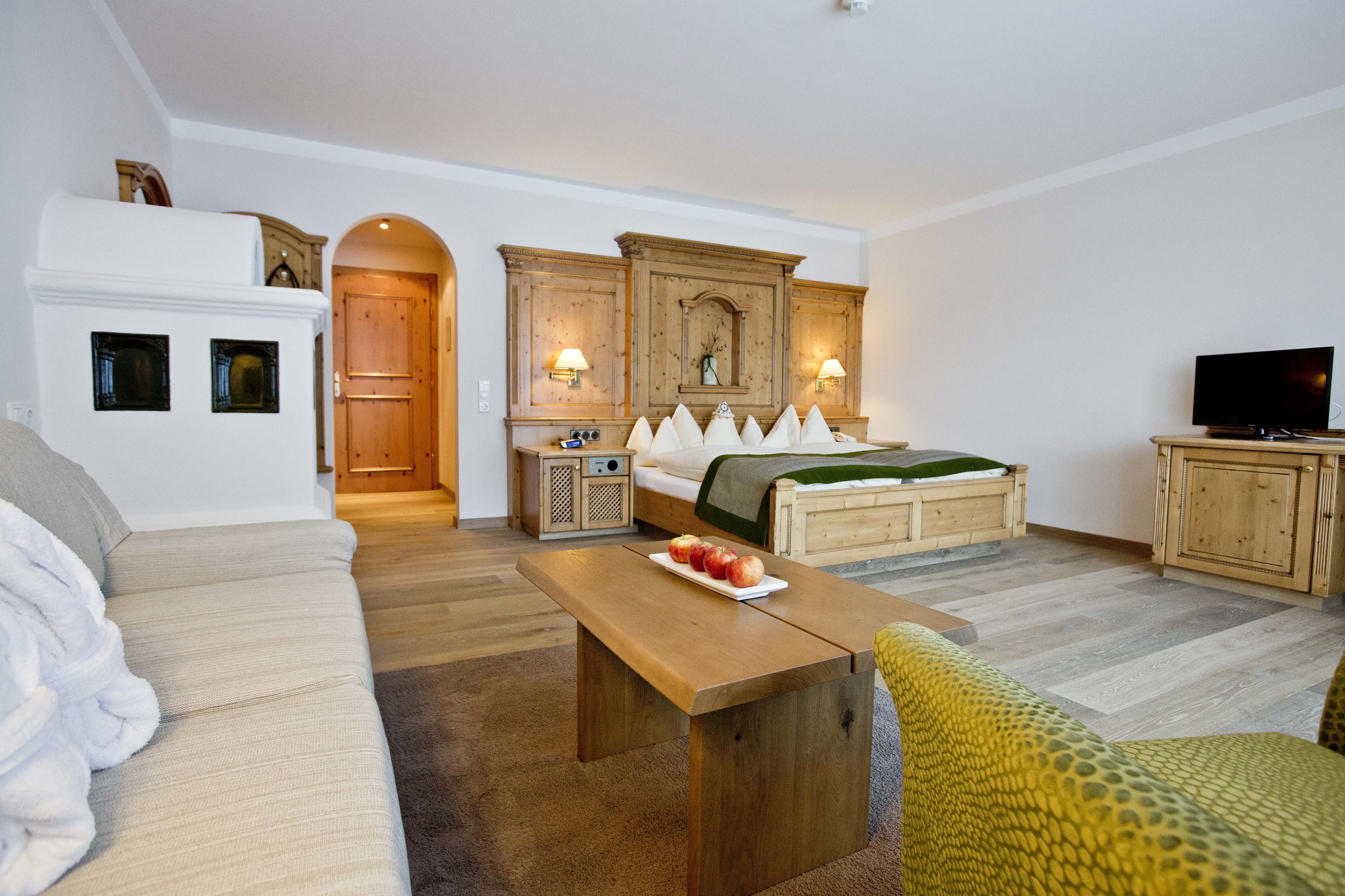 A cozy living room with a sofa, a wooden coffee table with apples, a bed with a green blanket, and a TV on a wooden cabinet.