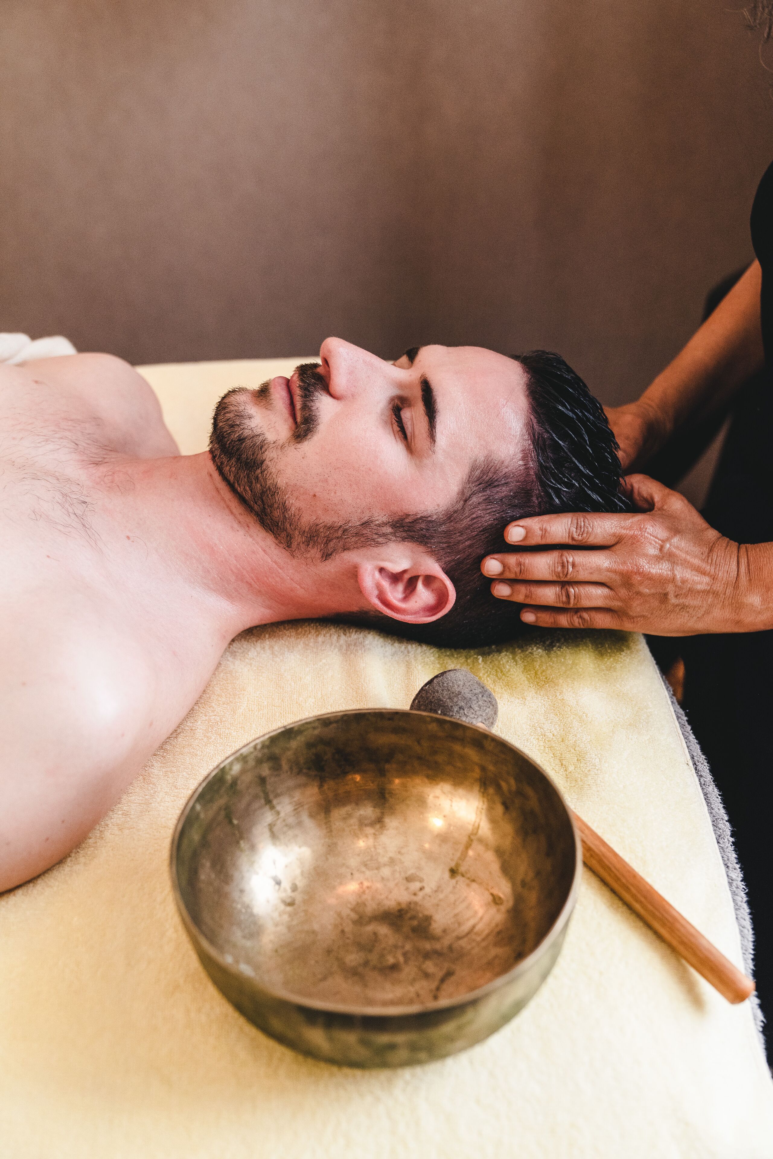 Eine Person erhält eine Kopfmassage mit einer Metallschüssel und einem Holzstab auf einem Handtuch.