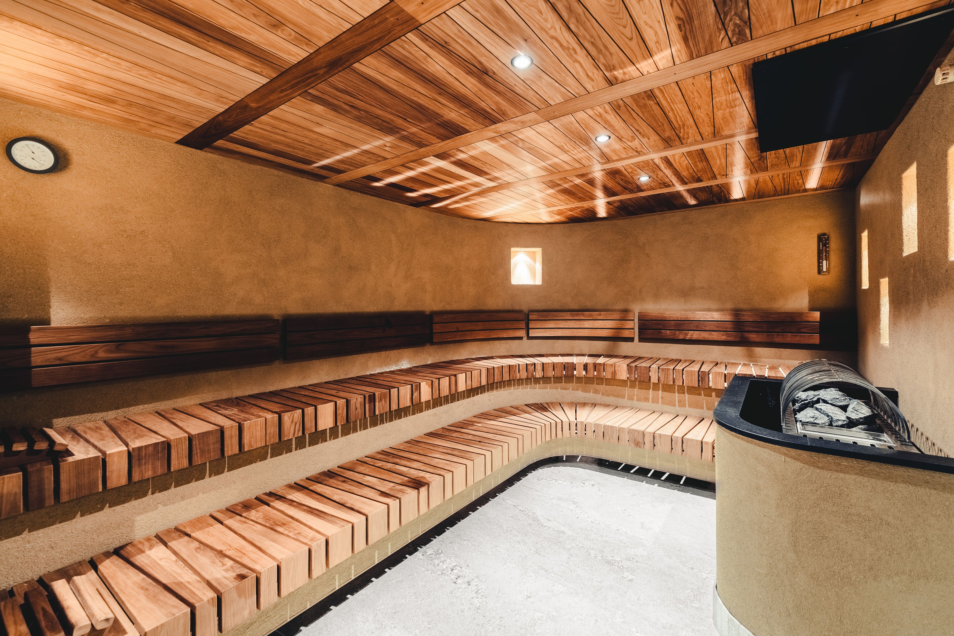 Uma sauna com bancos de madeira dispostos em formato semicircular, um relógio na parede e uma pequena janela para iluminação natural.