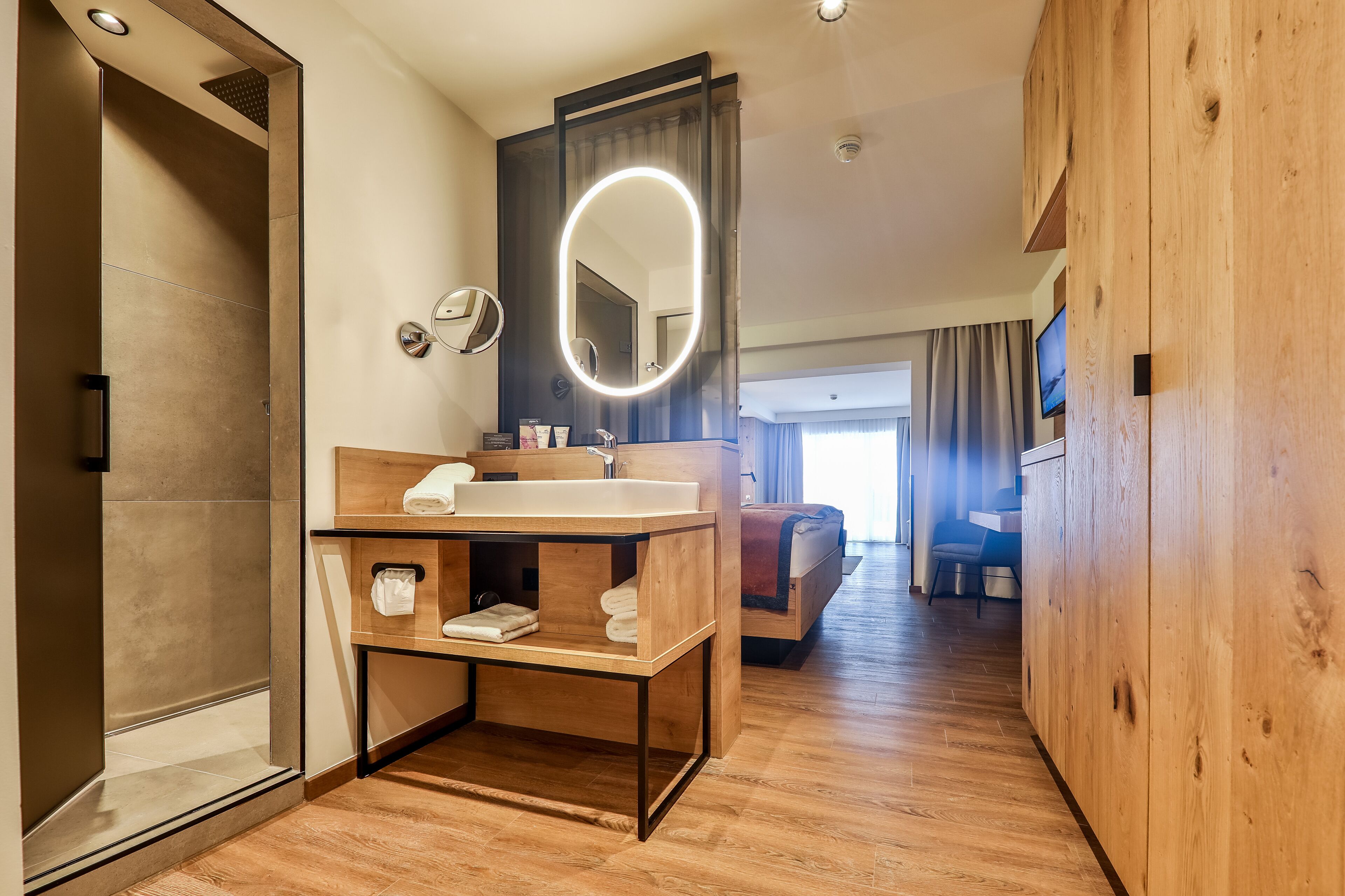 Un bagno moderno con un mobile vanitario in legno, un grande specchio con illuminazione a luce soffusa e una cabina doccia, con vista sul soggiorno.