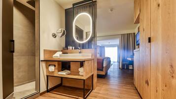 Un bagno moderno con un mobile vanitario in legno, un grande specchio con illuminazione a luce soffusa e una cabina doccia, con vista sul soggiorno.
