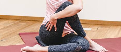 A person sitting on a red exercise mat in a room with wooden flooring and a plain wall.