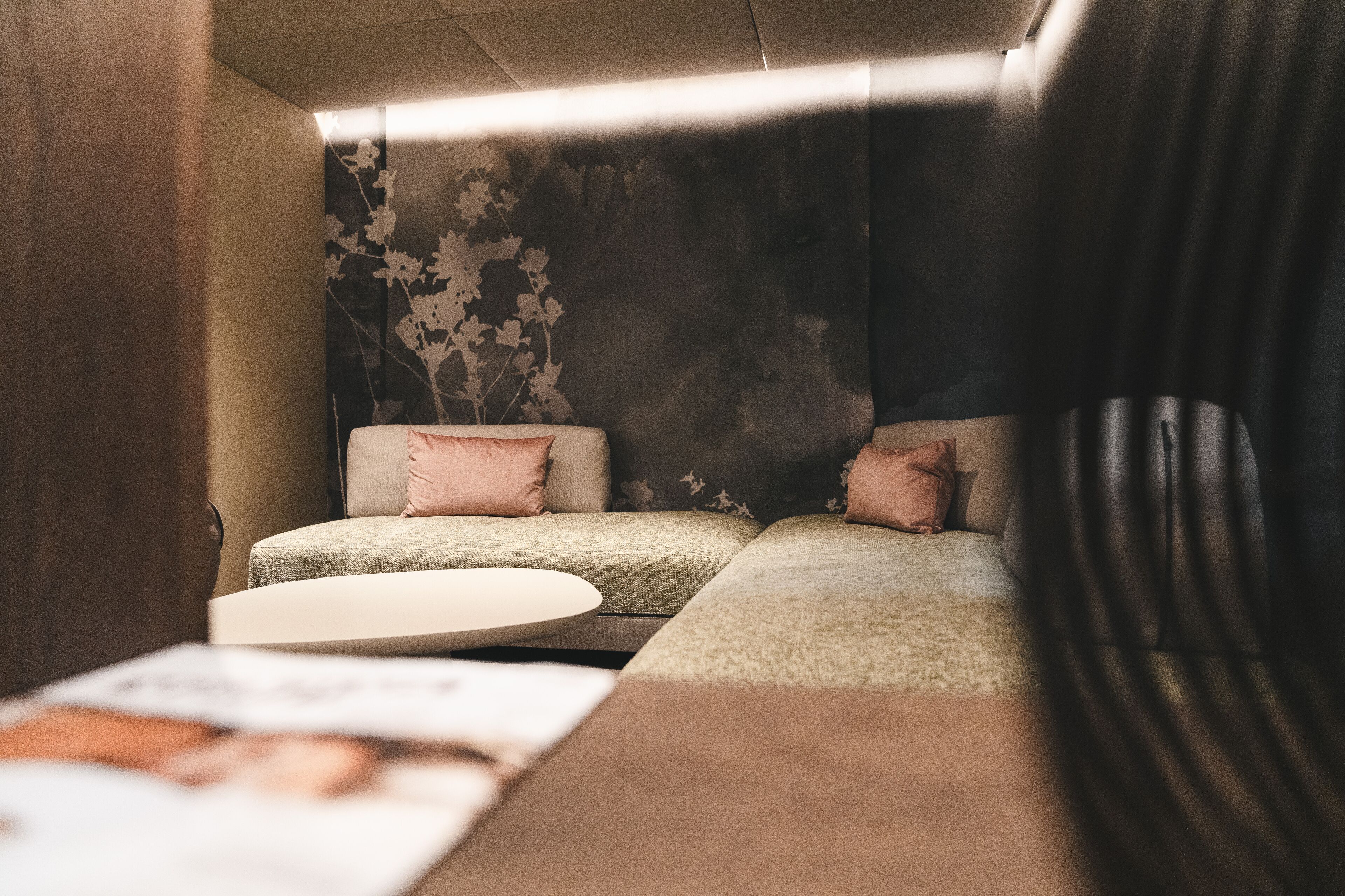 A cozy seating area with two cushioned sofas and a round table, featuring a dark wall with a floral pattern and a window with curtains.