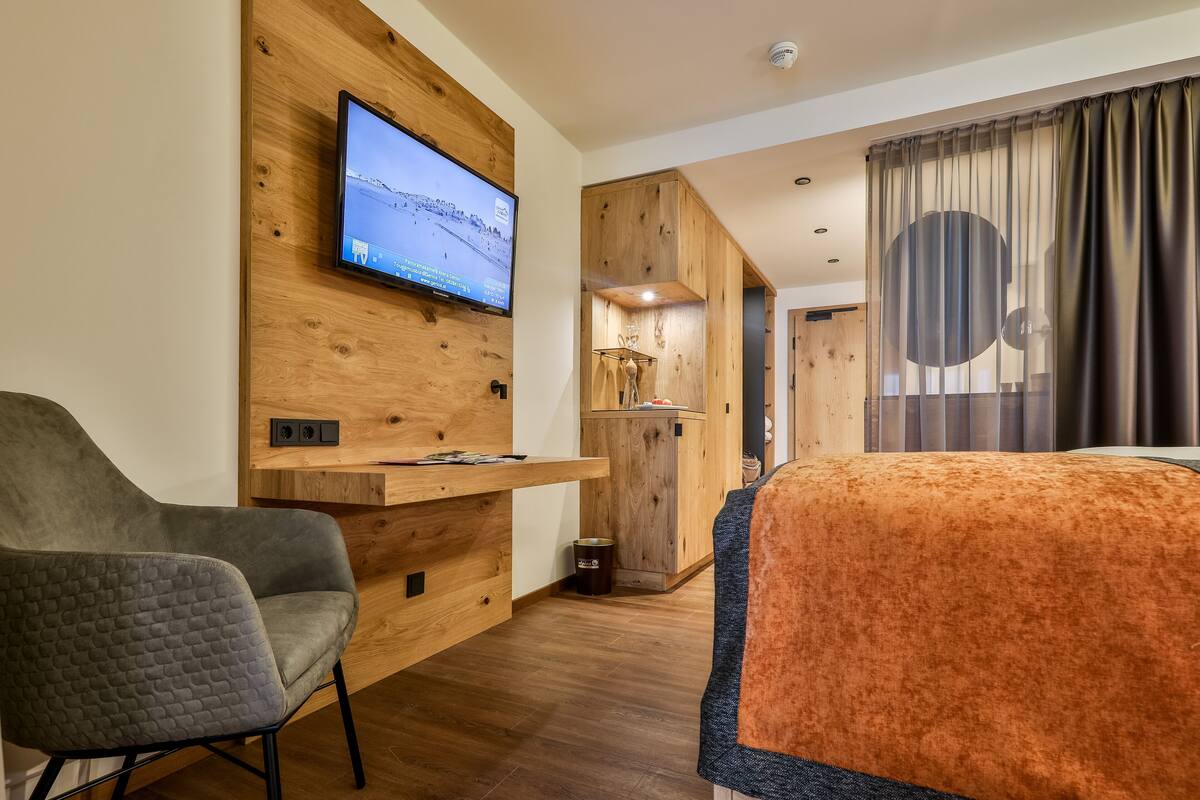 A modern hotel room with a flat-screen TV mounted on a wooden wall, a grey upholstered chair, a wooden cabinet with a small table, and a large bed with an orange blanket.