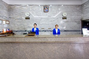 A reception desk with two staff members in blue uniforms, a marble wall with clocks, and a framed picture above the clocks. - Hat Yai Rama Hotel (Hat Yai)