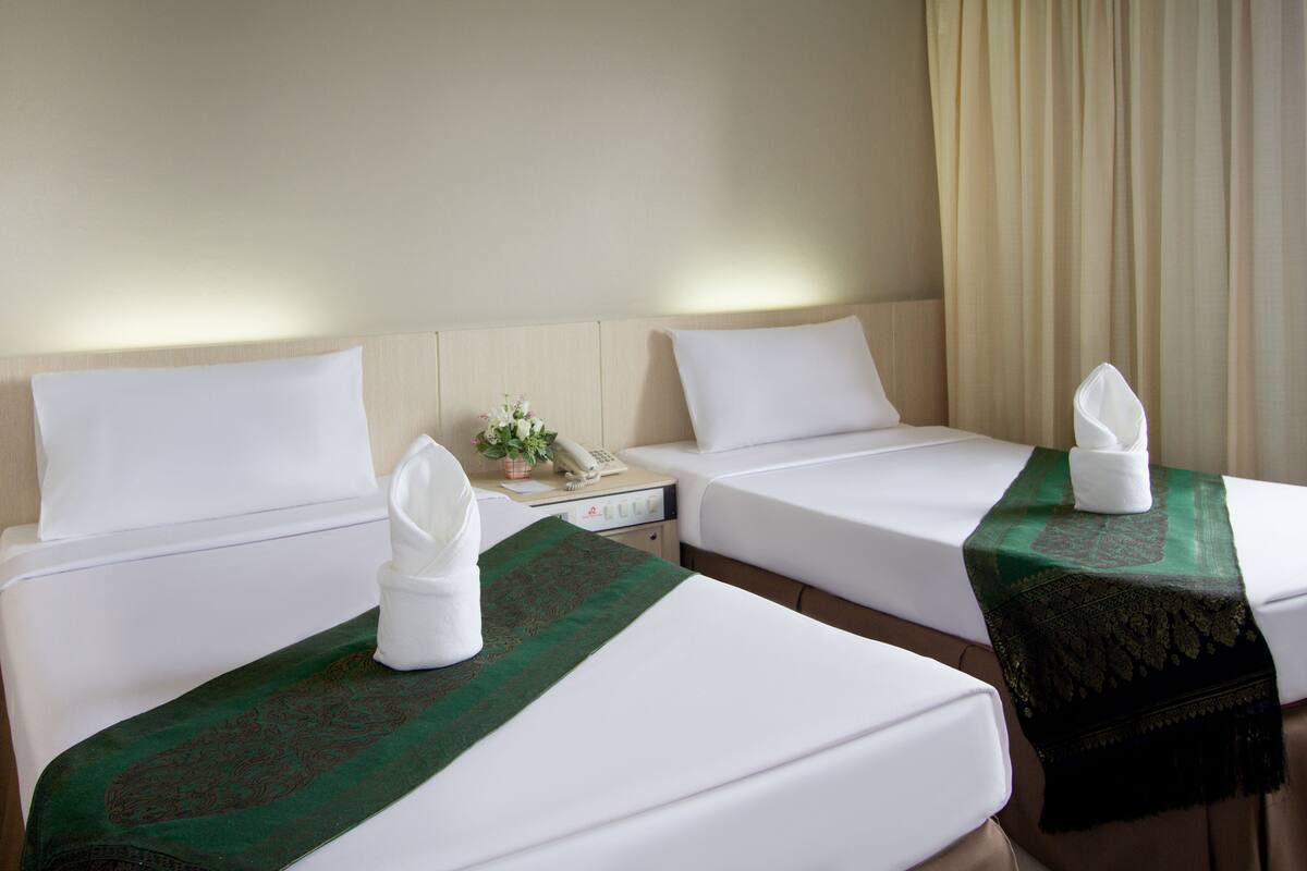 A hotel room with two beds, each with white linens and a green decorative runner. There is a bedside table with a telephone and a small plant.