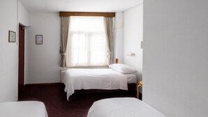 Una habitación de hotel con dos camas individuales, un ventanal con cortinas, un cabecero de madera y una mesita de noche pequeña.