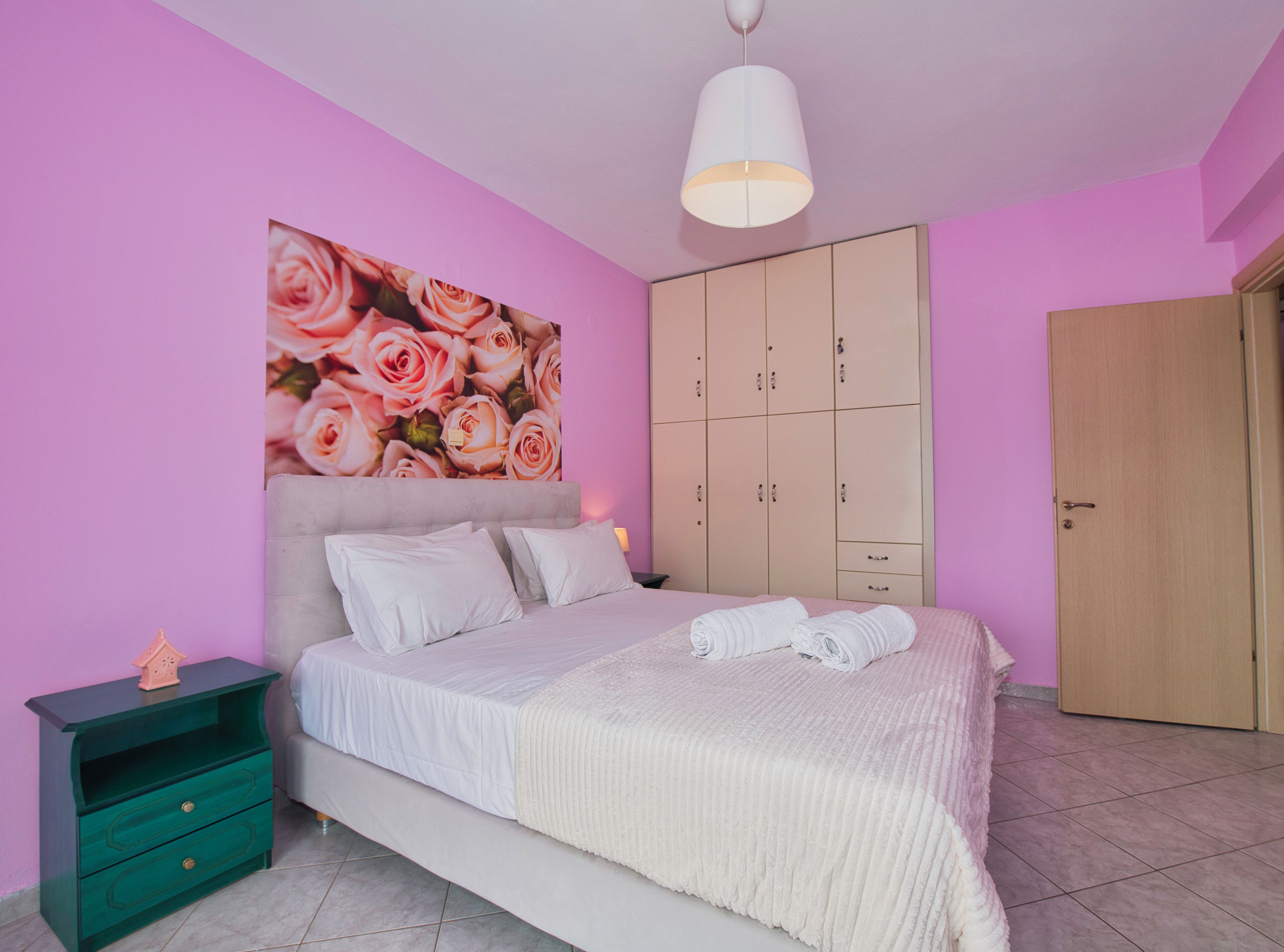 A bedroom with a bed, a nightstand, a wardrobe, and a pink wall with a floral mural.