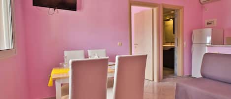 A pink-walled room with a dining table, chairs, a sofa, a TV, and a refrigerator.