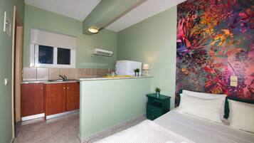Habitación de hotel pequeña con kitchenette, una cama con ropa blanca, un mural floral colorido y una mesita de noche verde.