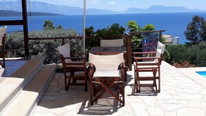 Um terraço com mesa e cadeiras, um guarda-sol branco e vista para o mar e montanhas.