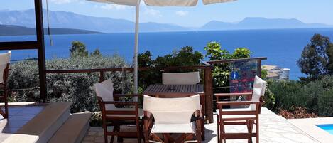 Une terrasse avec une table et des chaises, un parasol blanc, et une vue sur la mer et les montagnes.