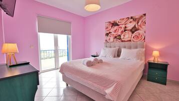 A bedroom with a large bed, a sliding glass door to a balcony, a painting of roses on the wall, and a green nightstand with a lamp.