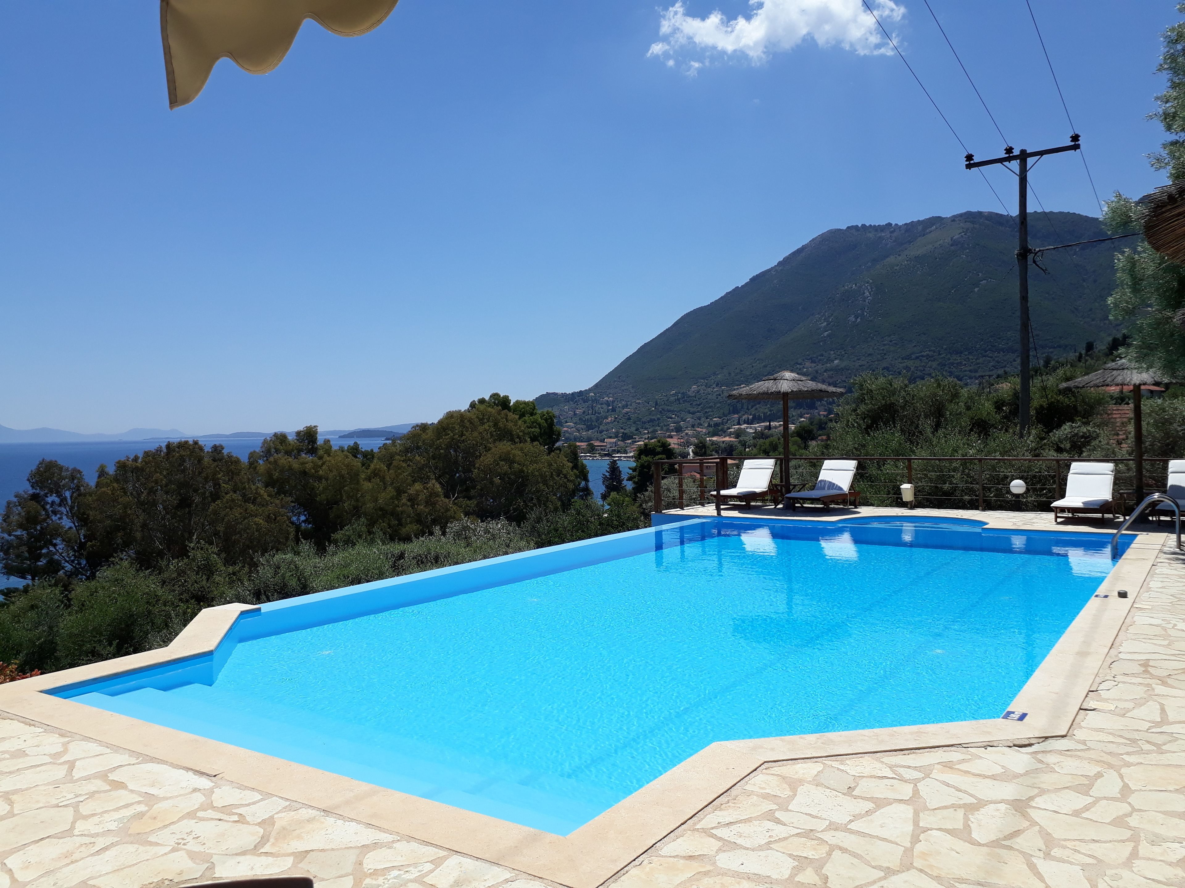 Una piscina con terraza de piedra, rodeada de sillones de descanso y vistas al mar y a las montañas.