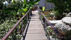 Uma trilha de madeira leva a um edifício com telhado de telhas, rodeado por vegetação exuberante e plantas floridas.