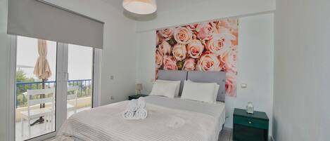 A bedroom with a large bed, a balcony with a chair, and a nightstand with a lamp.