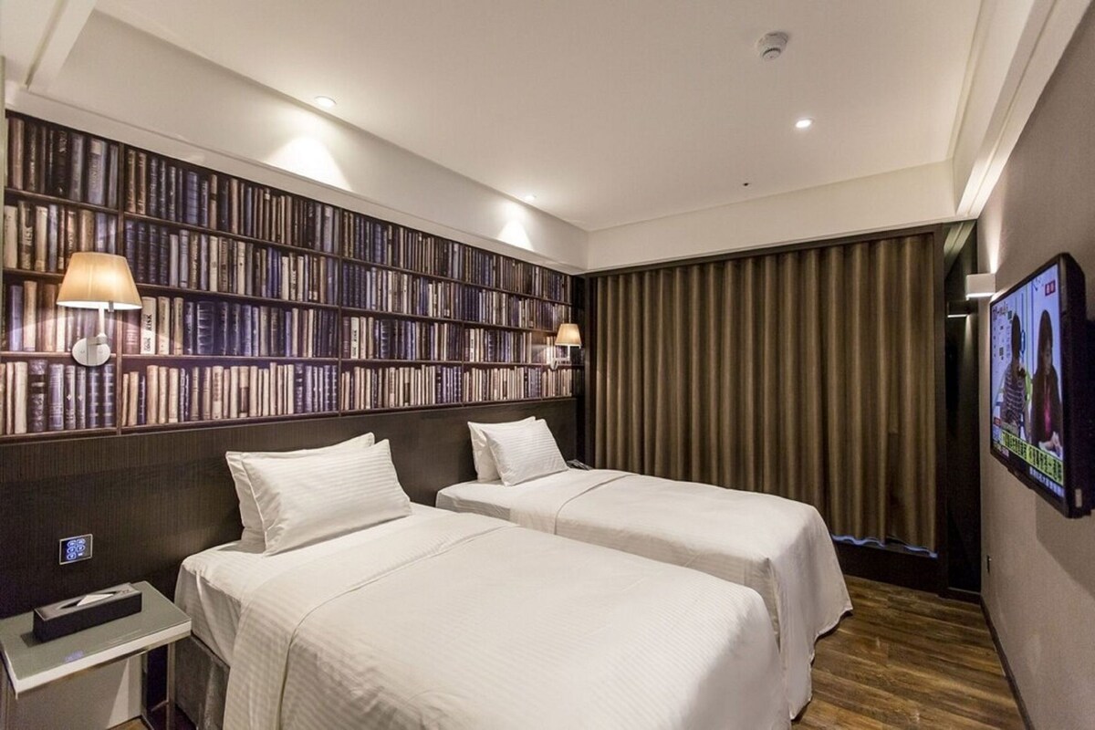 A hotel room with two beds, a bookshelf, a television, and a small table.