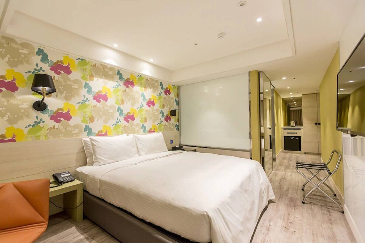 Quarto de hotel com cama, mesa de cabeceira, luminária, cadeira, espelho e banheiro visível por uma porta aberta.