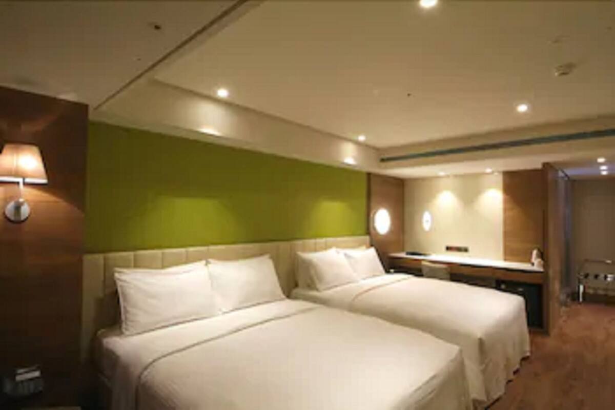Quarto de hotel com duas camas, uma parede de destaque verde e uma pequena cozinha compacta.