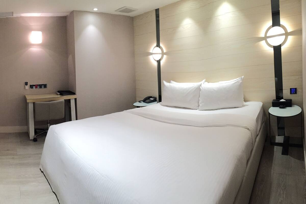 Quarto de hotel com cama, mesas de cabeceira, escrivaninha, televisão de tela plana e luminária fixada na parede.