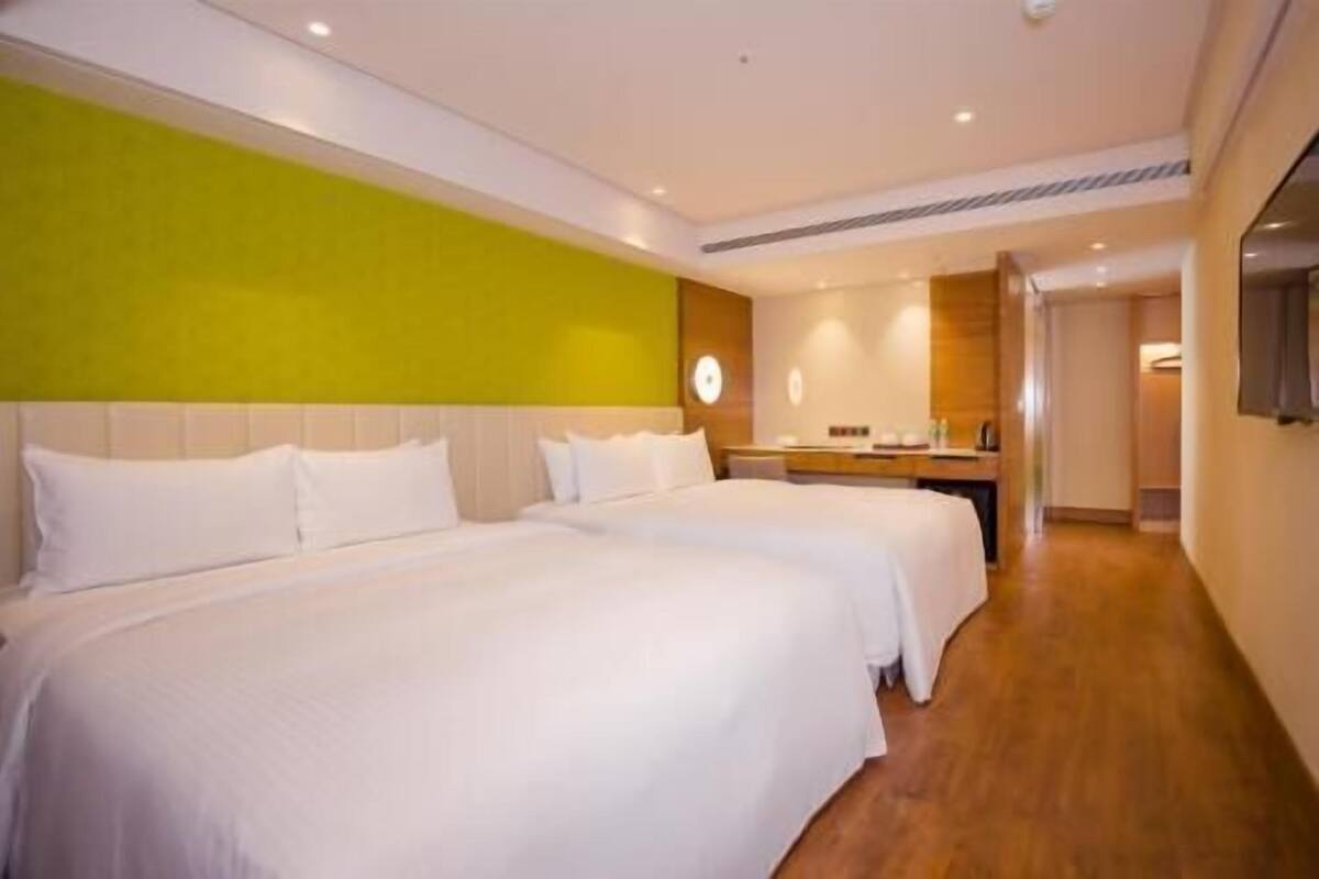 Quarto de hotel com duas camas, piso de madeira, parede verde e uma pequena cozinha compacta.