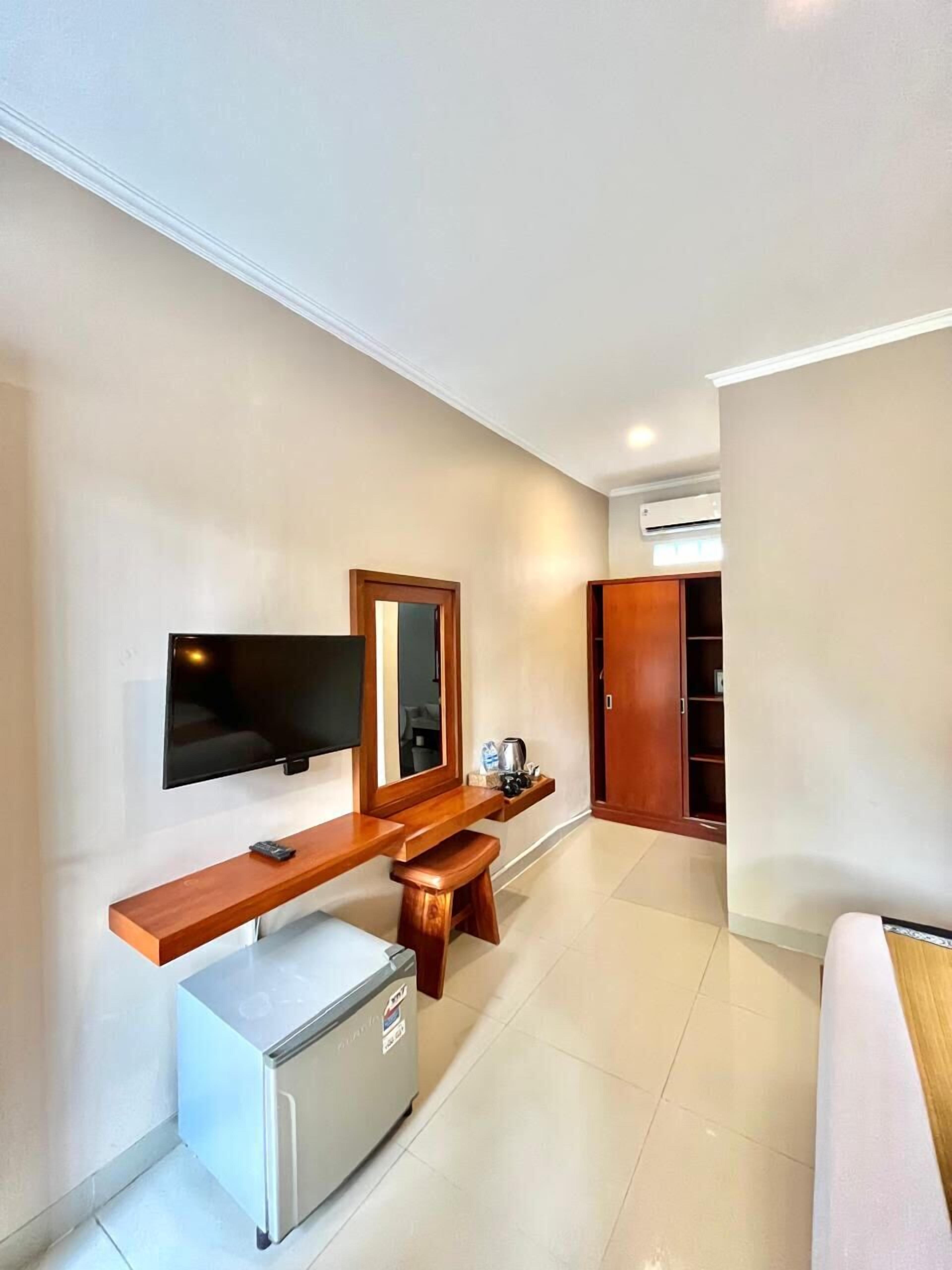 A modern hotel room with a flat-screen TV mounted on the wall, a wooden bench with a small stool, a mini-fridge, and a wardrobe.
