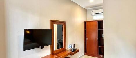 A modern hotel room with a flat-screen TV mounted on the wall, a wooden bench with a small stool, a mini-fridge, and a wardrobe.